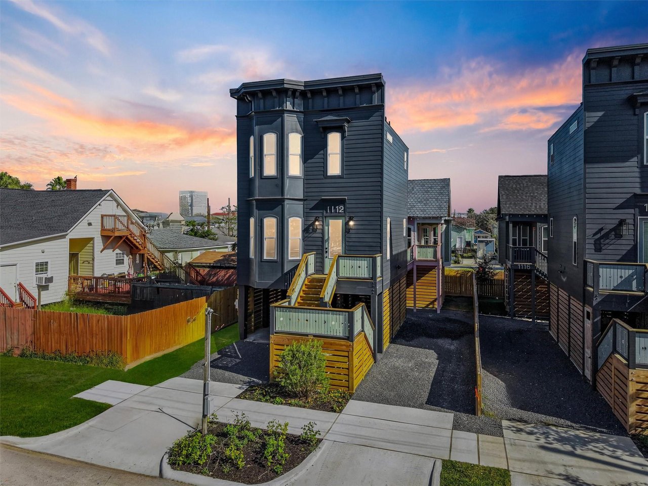 Dark two-story elevated tiny home with yellow stairs and a deck among neighboring houses at sunset.
