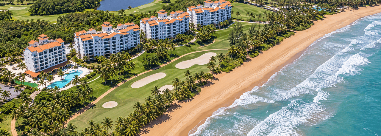 Oceanfront condominium complex with orange tile roofs, a golf course, pool, and palm-lined beach.