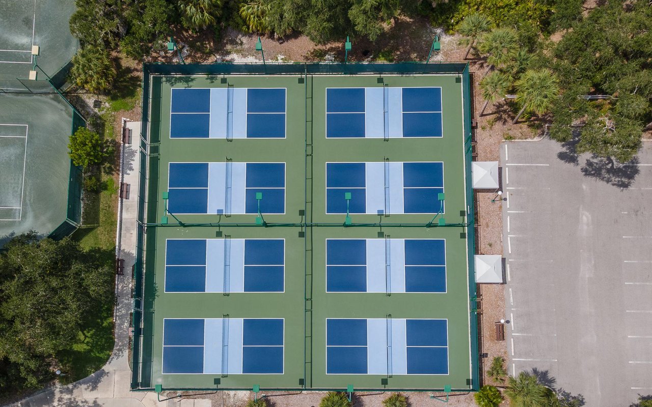 Fenced pickleball courts with blue playing surfaces and a parking lot alongside.