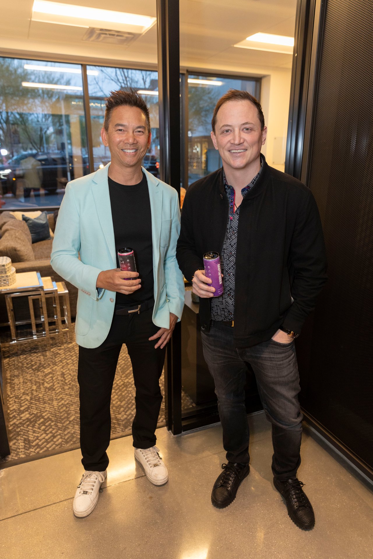 Two men pose with drinks at an indoor event, one wearing a light blue blazer and the other a black jacket.
