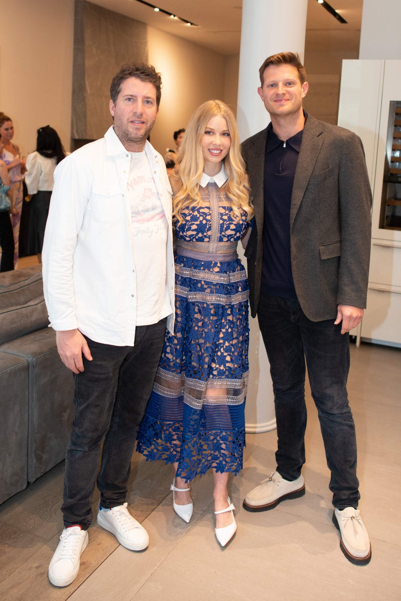 Three people pose together at an indoor event; the woman wears a blue lace dress.