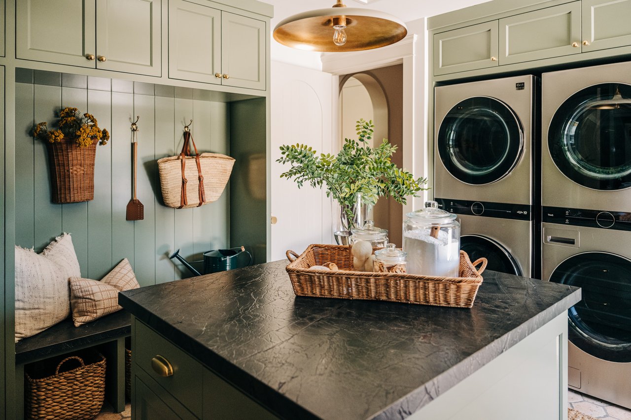 Laundry room with sage-green cabinets, stacked washer and dryer, built-in bench, and dark stone island.