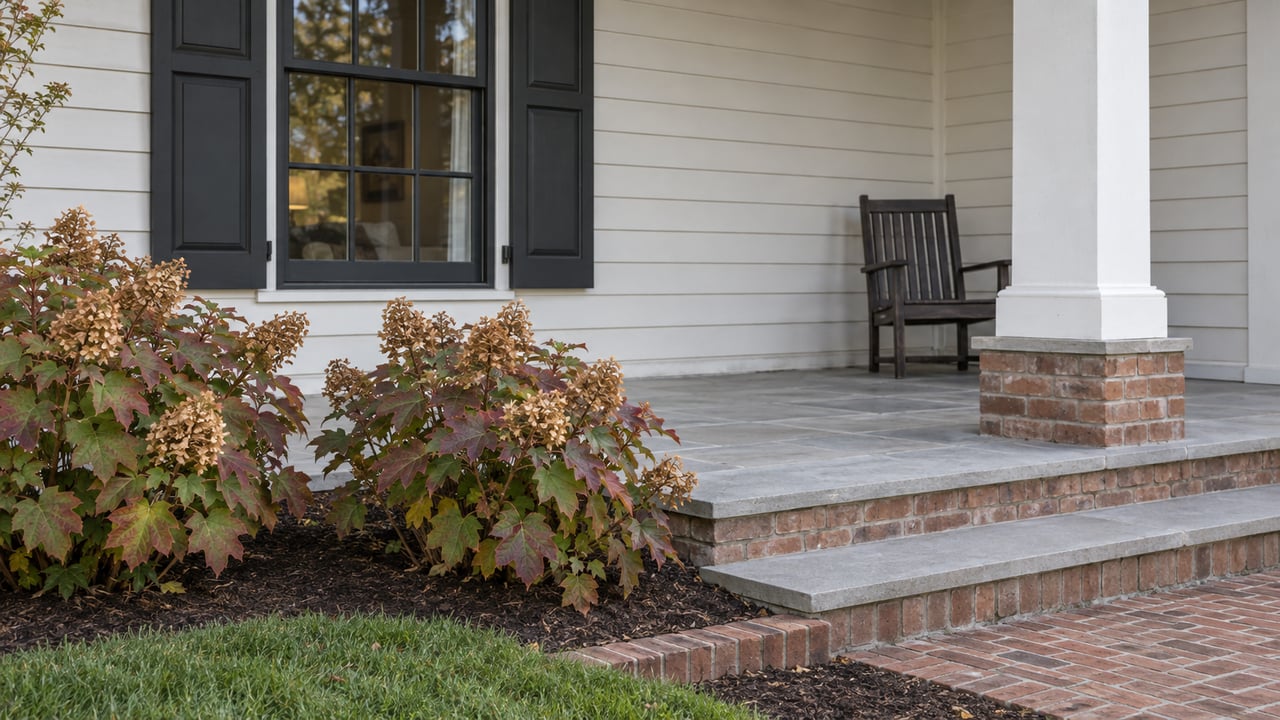 White-sided home with black shutters, a limestone porch, brick steps and oakleaf hydrangeas turning burgundy.