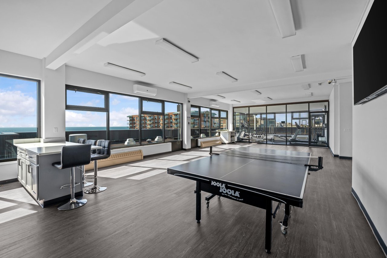 Communal recreation room with a ping-pong table, exercise equipment, and ocean views through large windows.