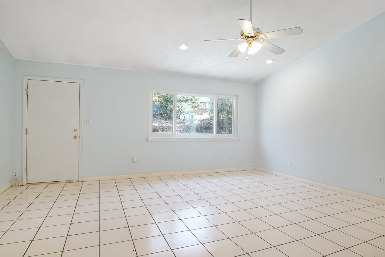 Empty room with pale blue walls, white tiled floor, and large window showing outdoor greenery. Ceiling fan with lights, simple and calm atmosphere.