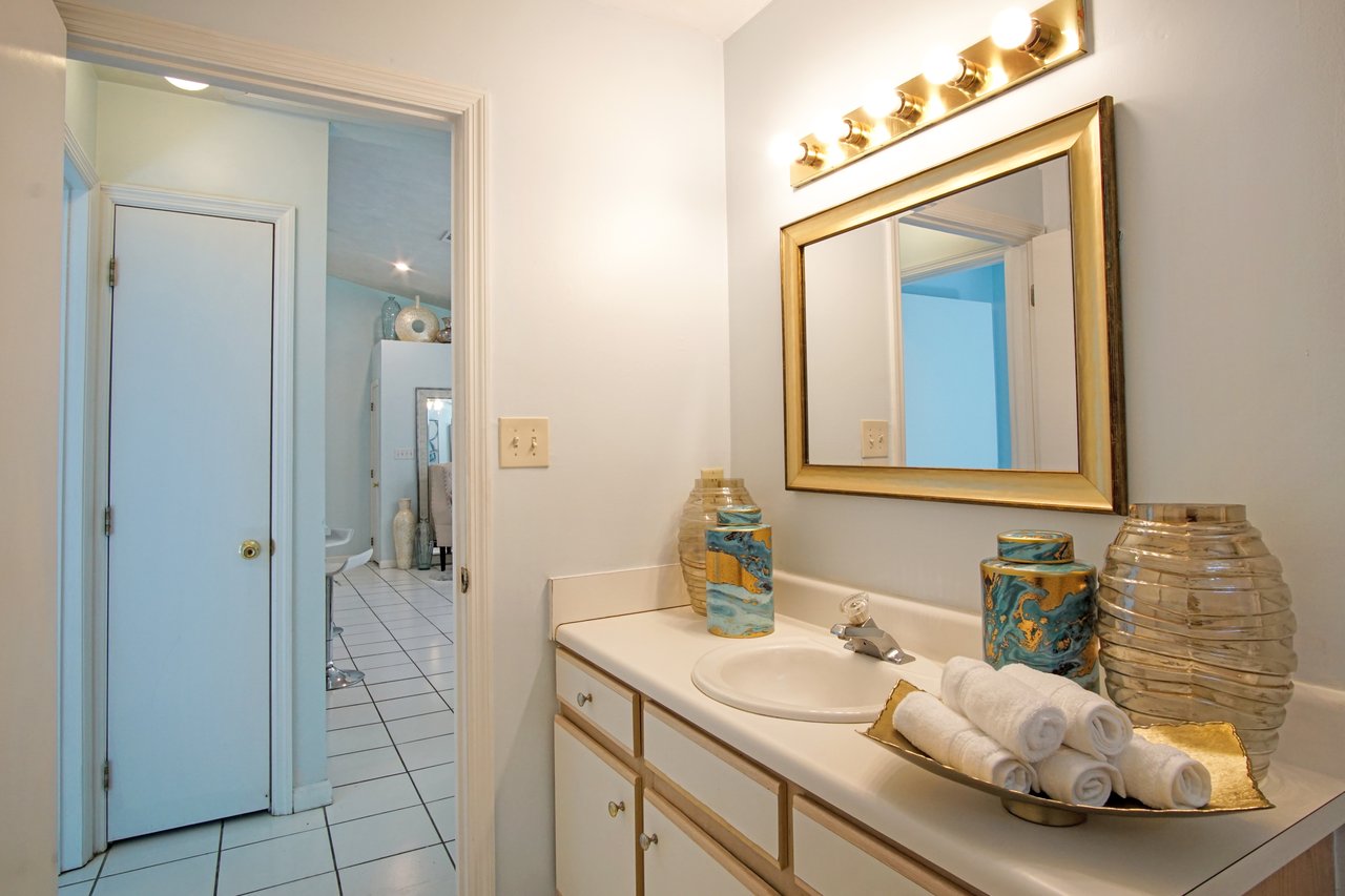 Bright bathroom with light blue walls and white tile floor. Features a large gold-framed mirror, countertop with rolled towels, decorative jars, and soft lighting.Bright bathroom with light blue walls and white tile floor. Features a large gold-framed mirror, countertop with rolled towels, decorative jars, and soft lighting.