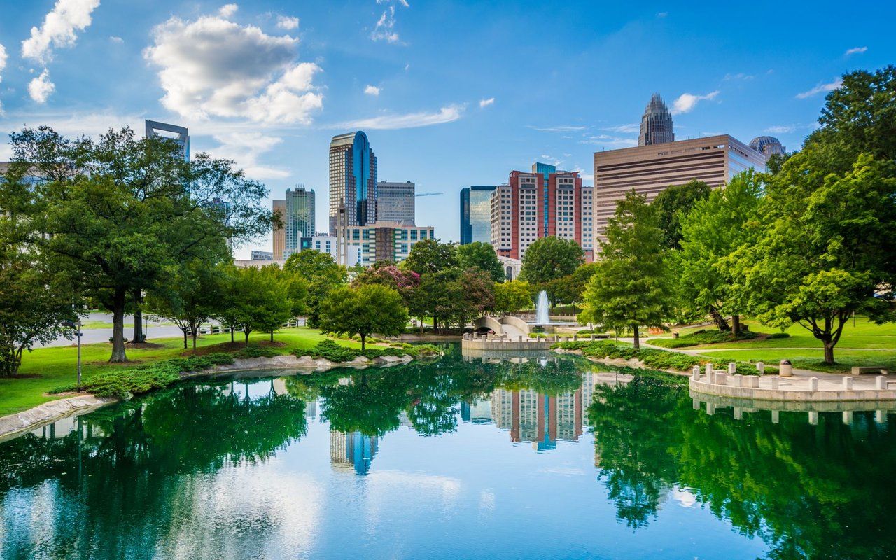 Uptown Charlotte skyline reflected in a park pond, with leafy trees and a fountain.
