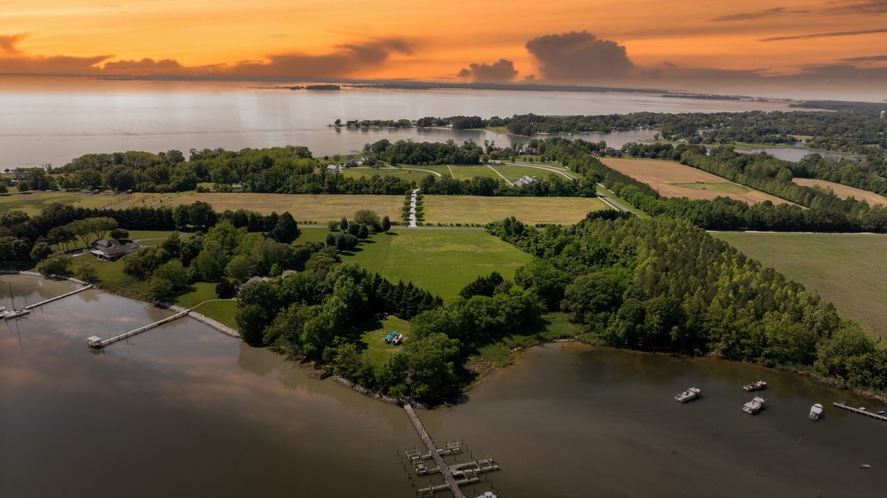 Aerial view of wooded waterfront acreage, fields, docks, and boats beneath an orange sunset.