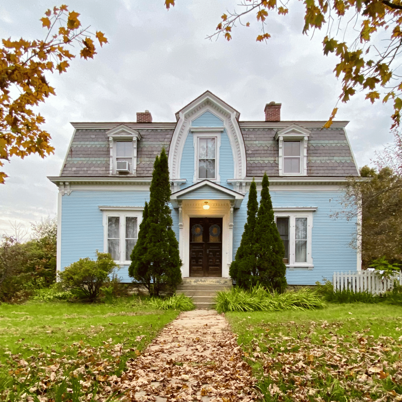 Light blue house with a mansard roof and arched dormer, framed by autumn leaves.