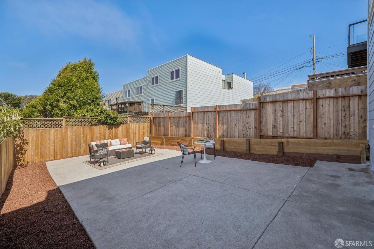 Fenced concrete patio with outdoor seating, mulched borders, and neighboring homes beyond the fence.