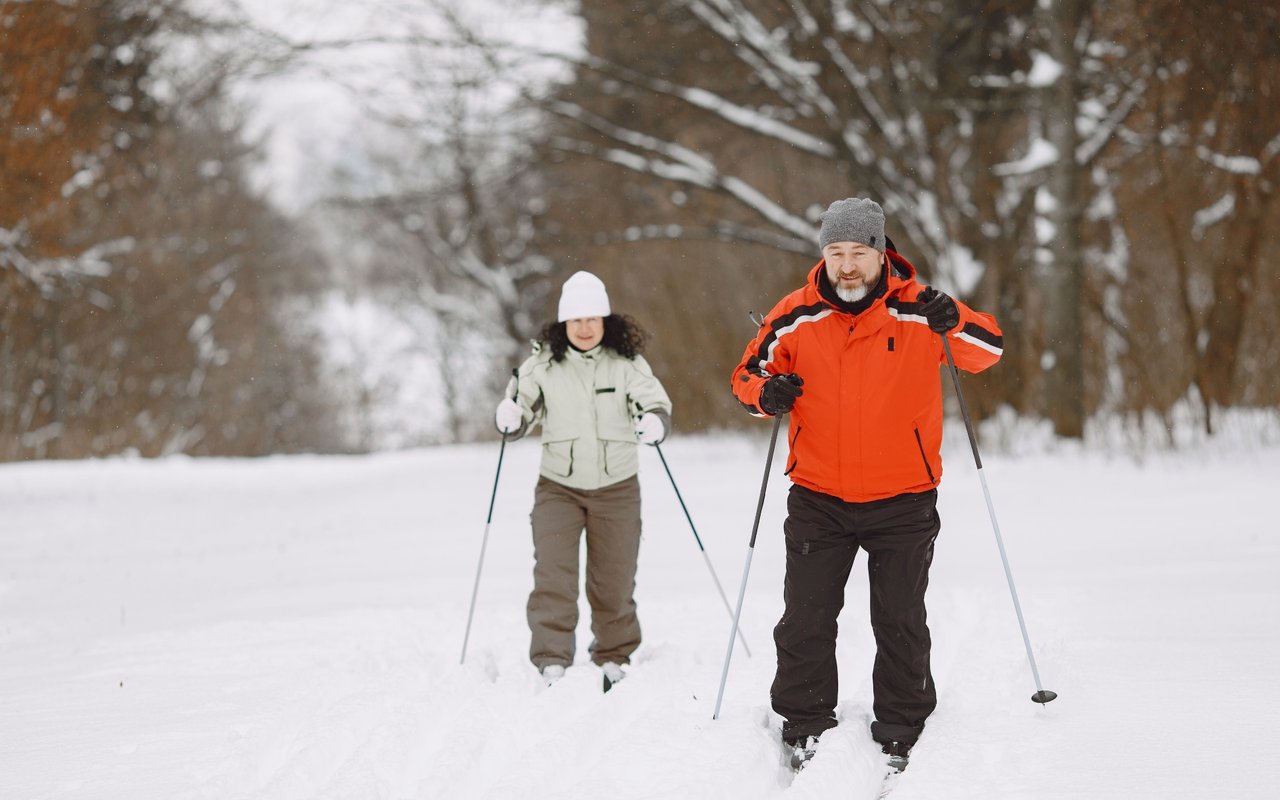 Two adults cross-country skiing through a snowy forest, the front skier wearing a bright orange jacket.