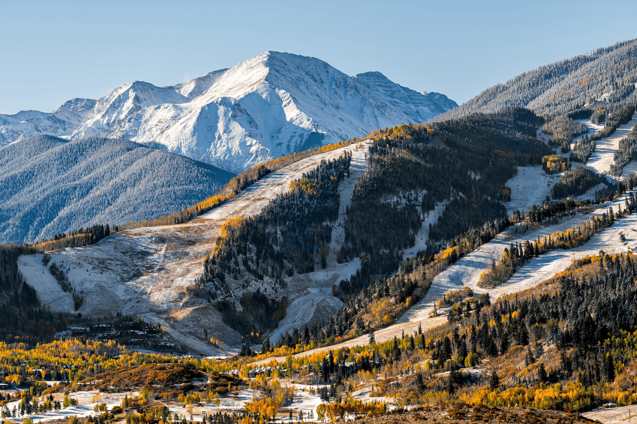 Aerial view of Aspen Highlands ski area during the seasonal transition, featuring golden aspen groves along the ski runs with snow-capped Mount Hayden dominating the background under clear blue skies.