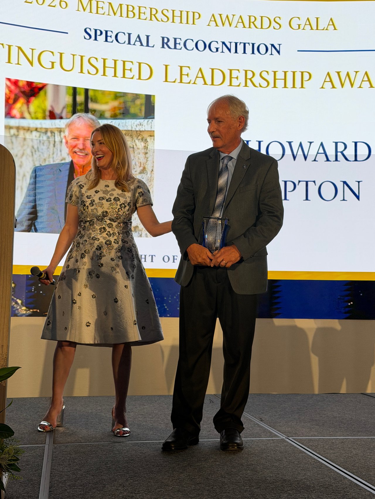 A woman with a microphone stands beside an older man holding a trophy at an awards gala.