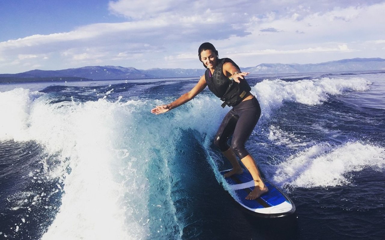 A woman wakesurfs on a blue board, balancing on a foamy wave with mountains in the distance.