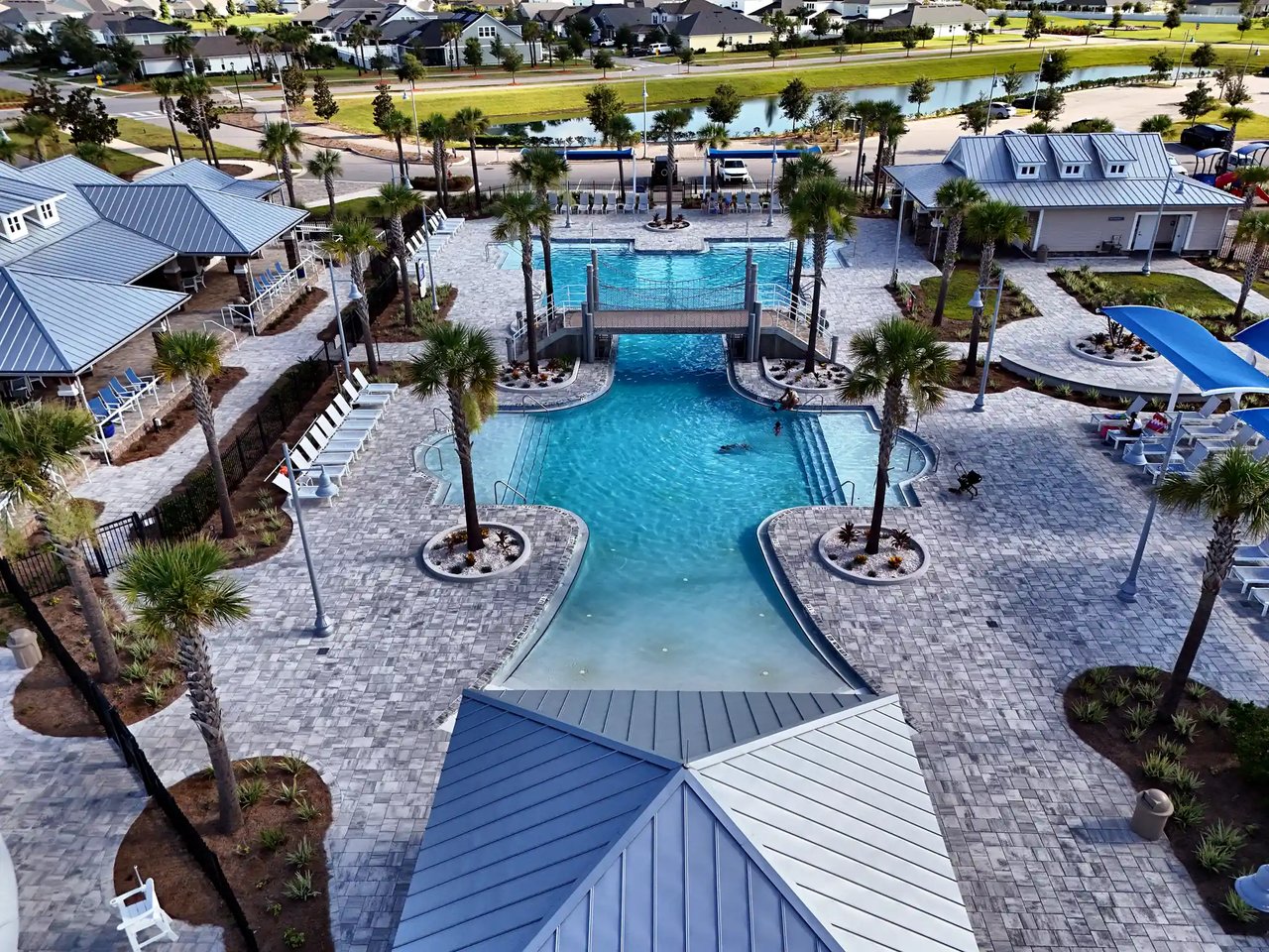 Aerial view of a community pool with a bridge, palm-lined stone deck, and nearby homes.