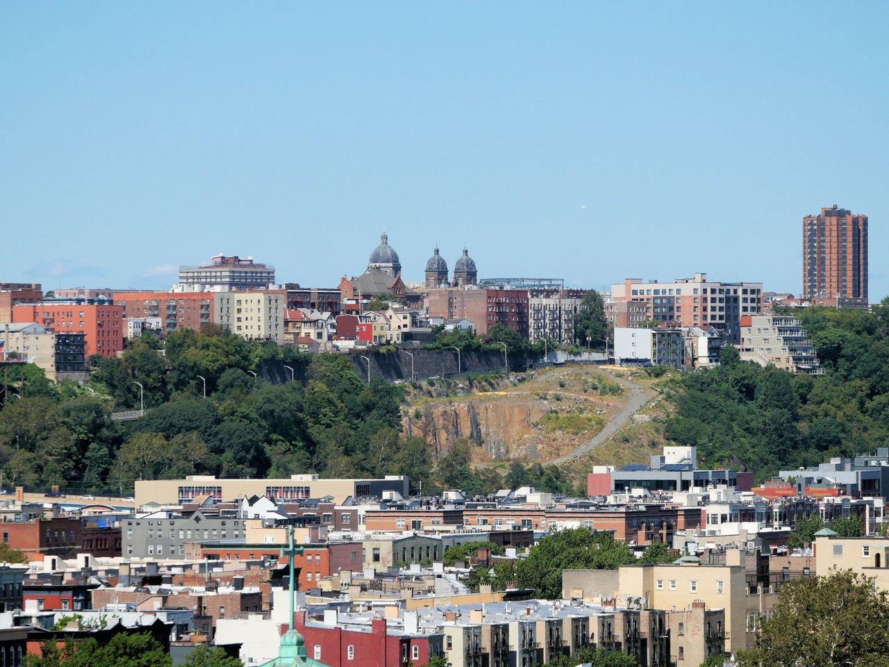 City buildings and church domes line a wooded rocky bluff beneath a clear blue sky.