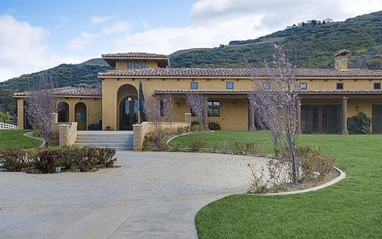 Tan stucco home with arched entryways and a curved driveway against a wooded hillside.