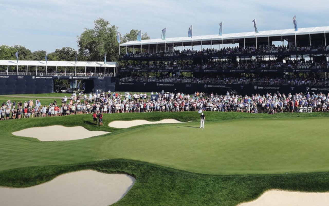 Golfer on a tournament course before packed grandstands, with sand bunkers in the foreground.