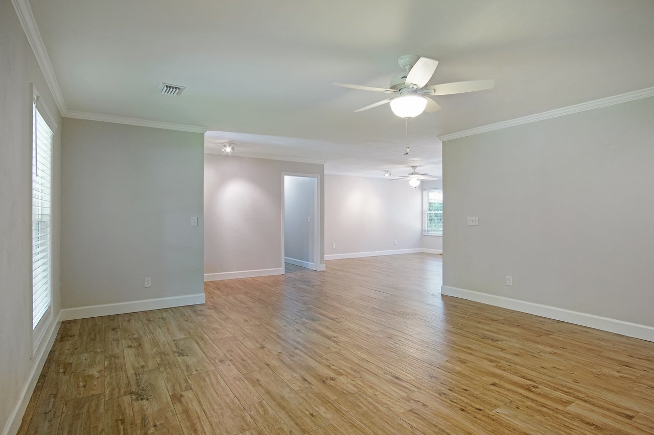 Spacious living area with wood flooring, white walls, and ceiling fans. Large windows provide natural light, creating a bright, inviting atmosphere.