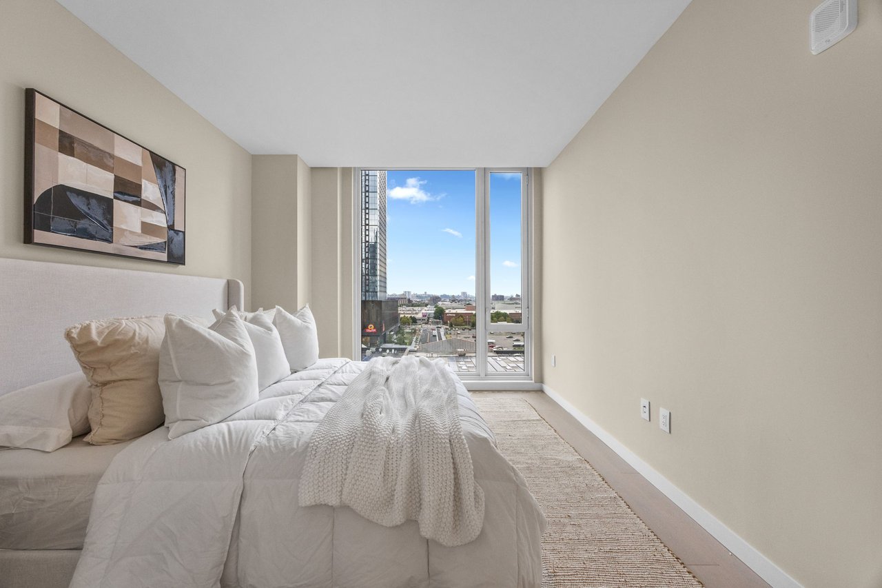 Staged bedroom with white bedding and a large window overlooking city buildings.