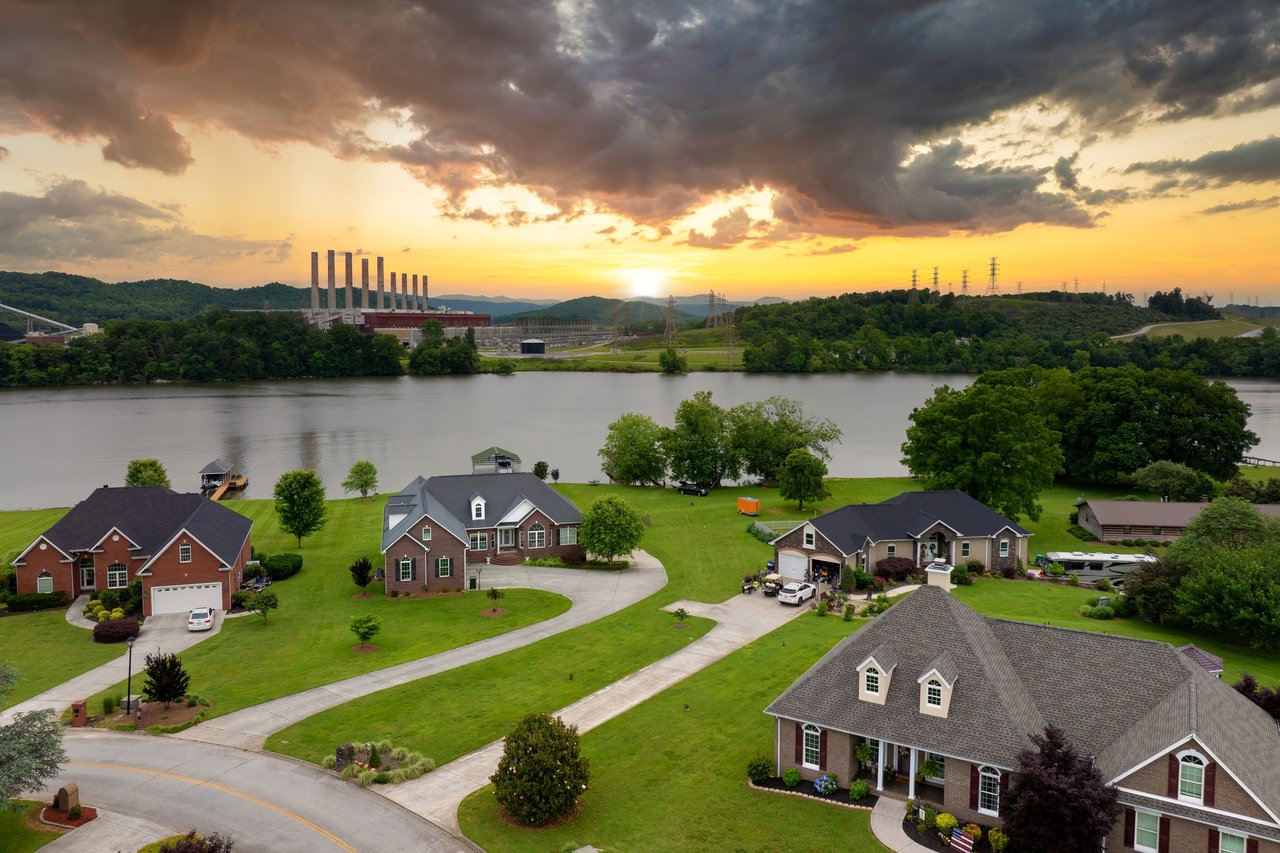 Riverfront homes and lawns beside a wide river, with wooded hills and a power plant beyond.
