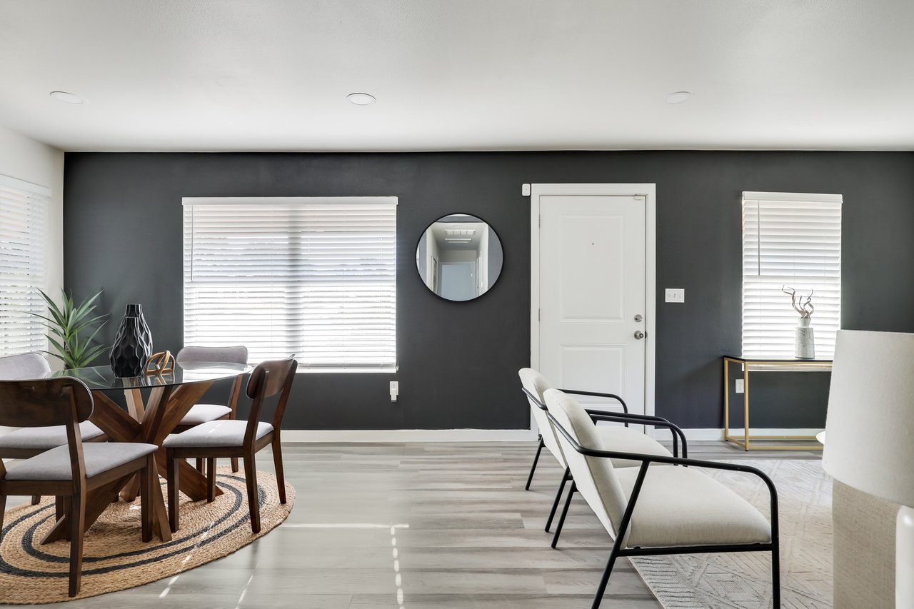 Furnished dining and sitting area with a charcoal accent wall, white front door, and large windows.
