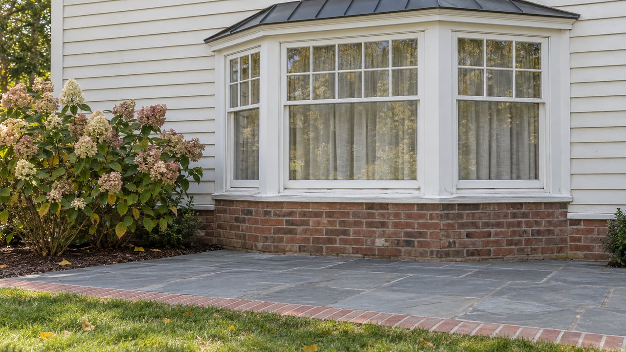 Home exterior with a large bay window, stone patio, and flowering shrubs.
