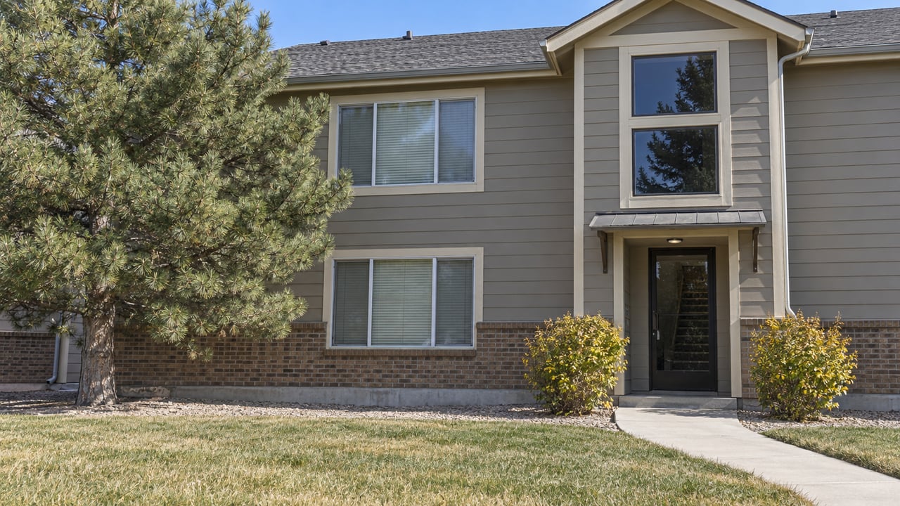 Two-story condominium facade with gray lap siding, brick base, shared glazed entrance and an autumn lawn beside a mature pine.