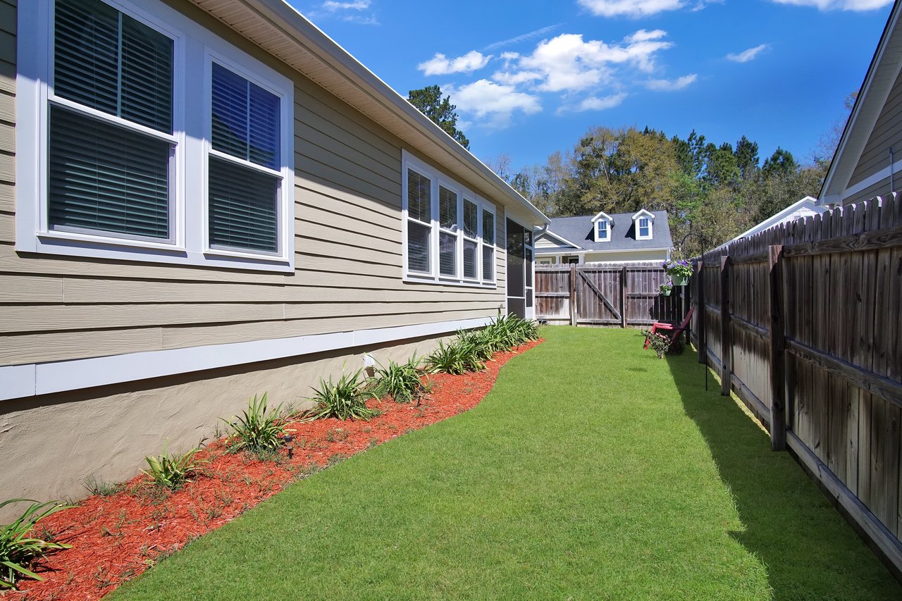 Side yard of a house with beige siding, bordered by a wooden fence. Lush green grass and red mulch beds line the house, under a clear blue sky.