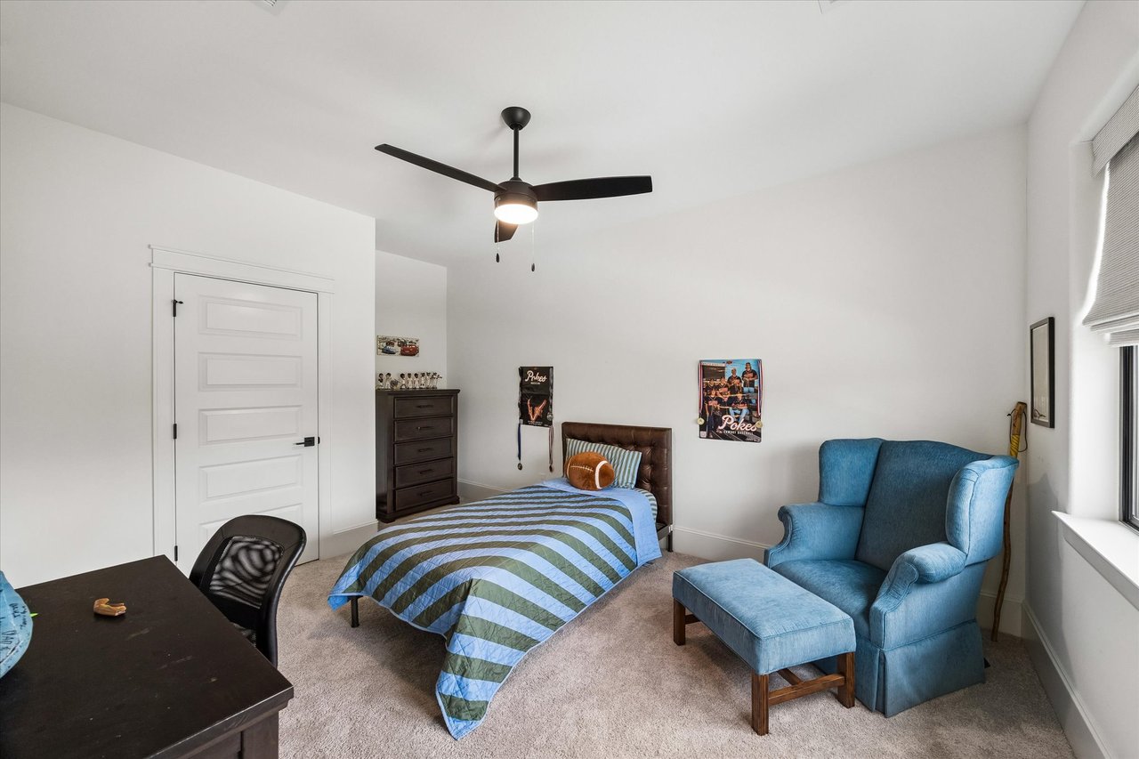 Bedroom with a blue-and-green striped bed, dark dresser, and blue armchair beside a window.