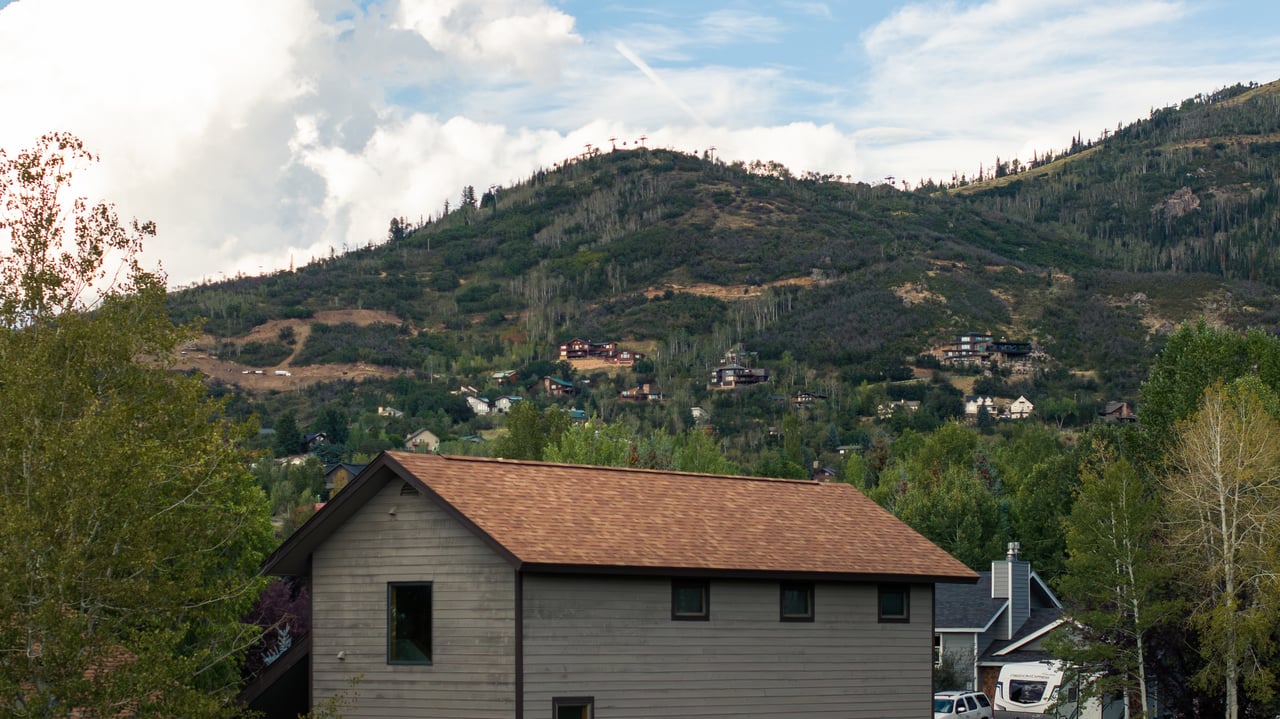 Gray-sided house with a brown roof below a forested mountain hillside.