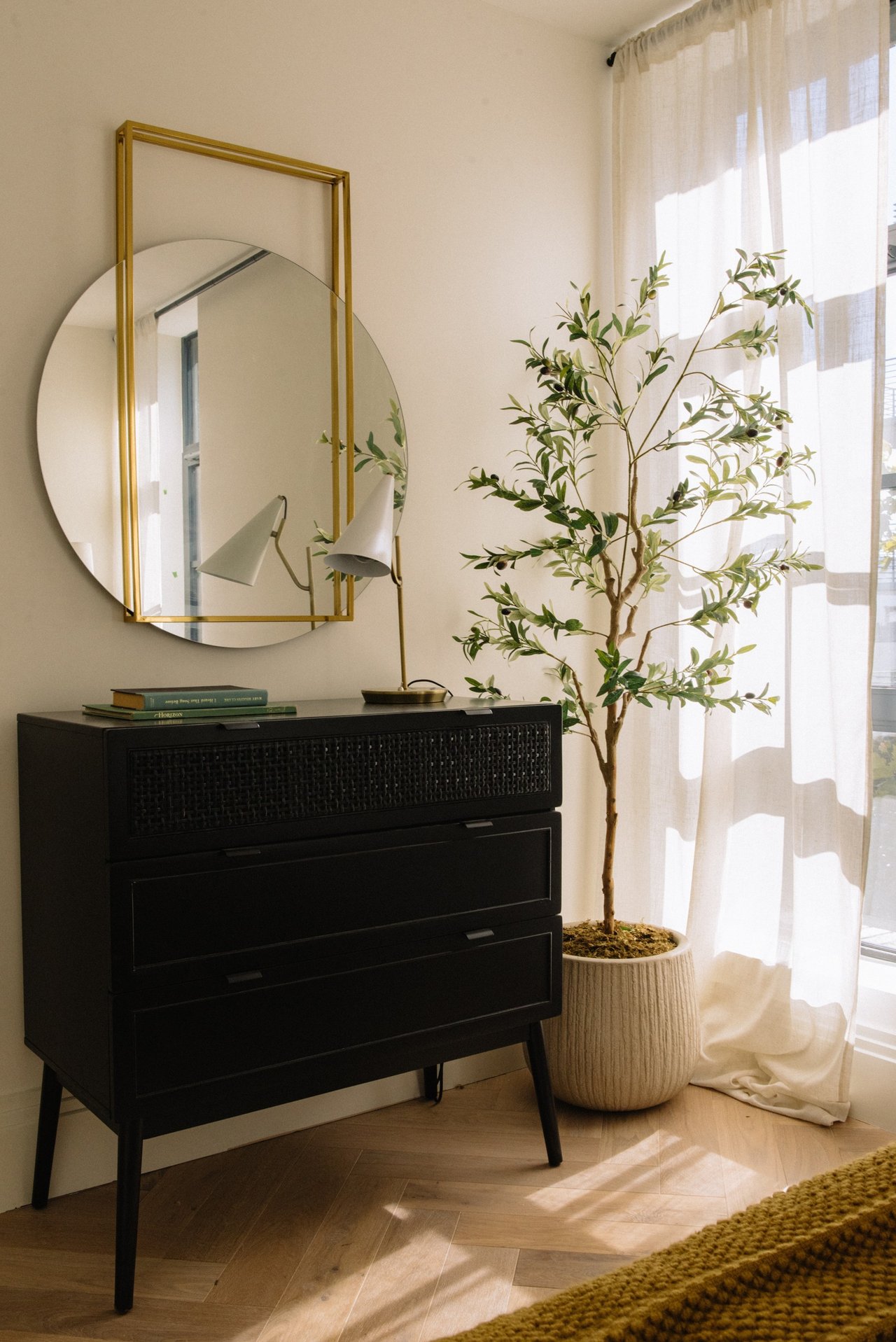 Bedroom corner with a black dresser, gold-framed round mirror, potted olive tree, and sheer curtains.