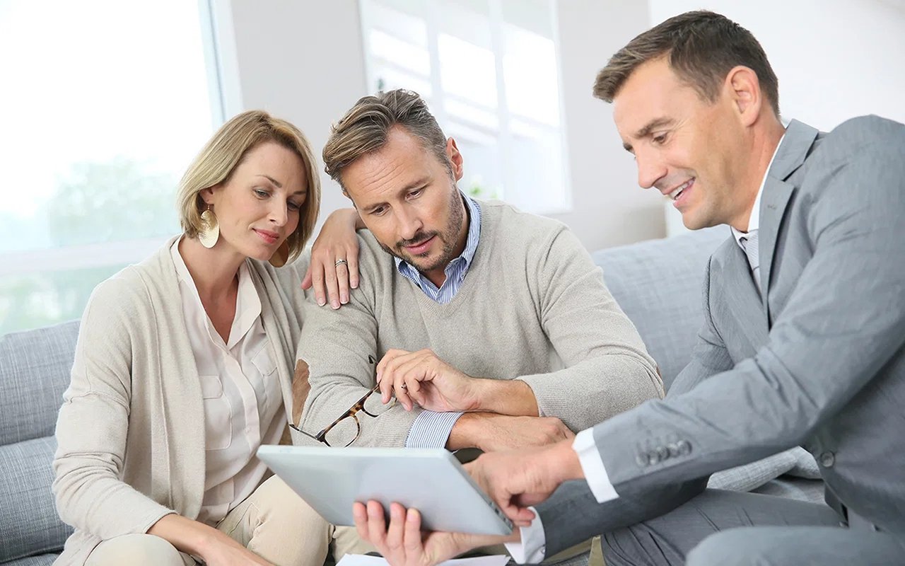 A real estate agent reviews a tablet with a couple seated beside him.