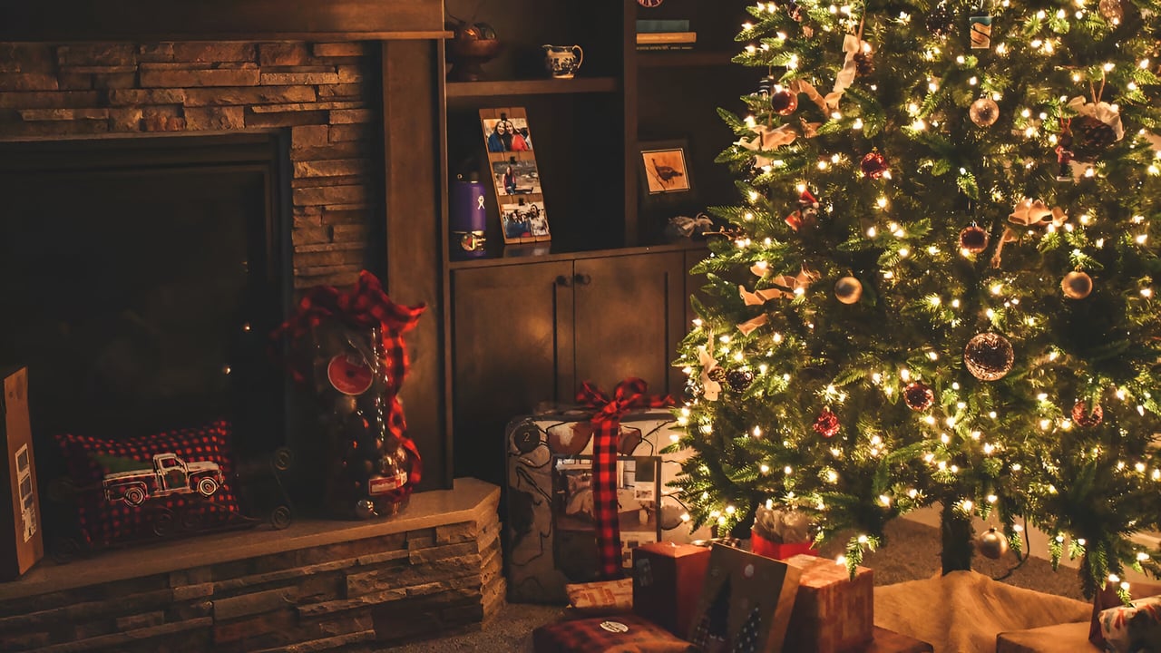 Holiday-decorated living room with a lit Christmas tree and presents beside a stone fireplace.