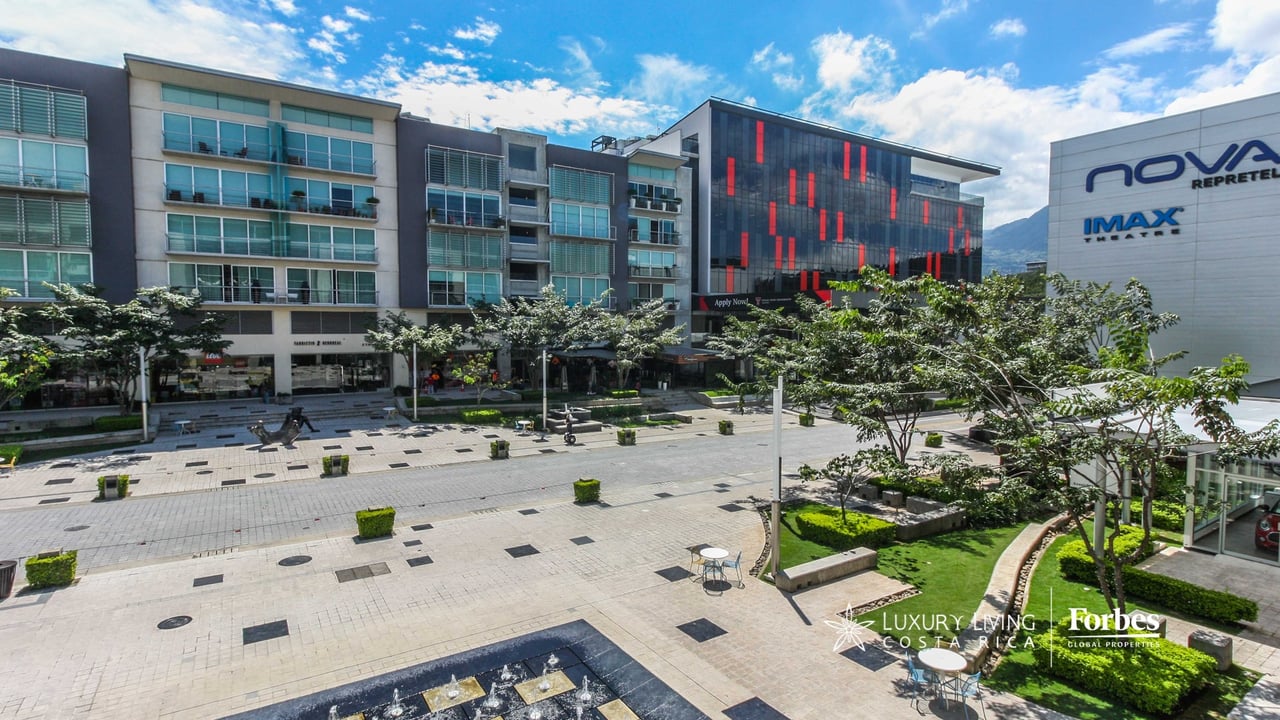 Modern Avenida Escazú complex with glass balconies overlooking a landscaped plaza and distant mountain.