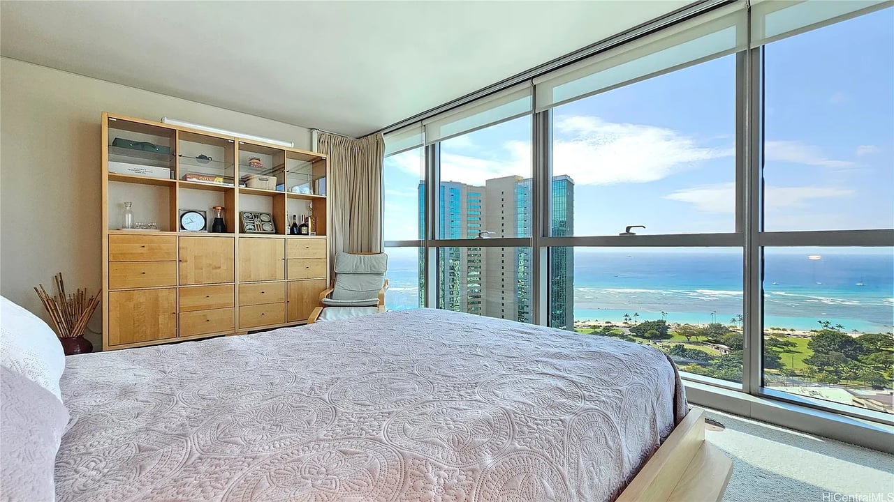 Furnished bedroom with floor-to-ceiling windows overlooking the ocean and nearby high-rise buildings.