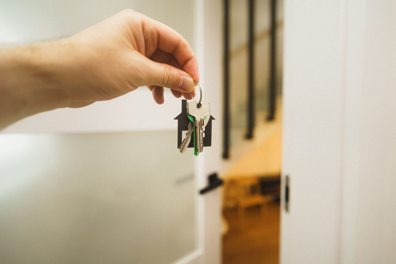 A hand holds house keys with a house-shaped keychain beside an open door.