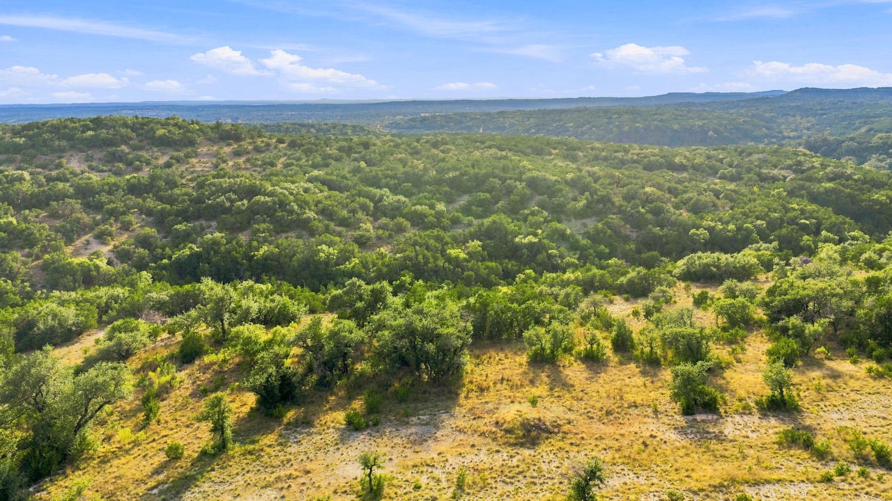 Aerial view of rolling ranchland with wooded hills, open grassland, and distant ridgelines.