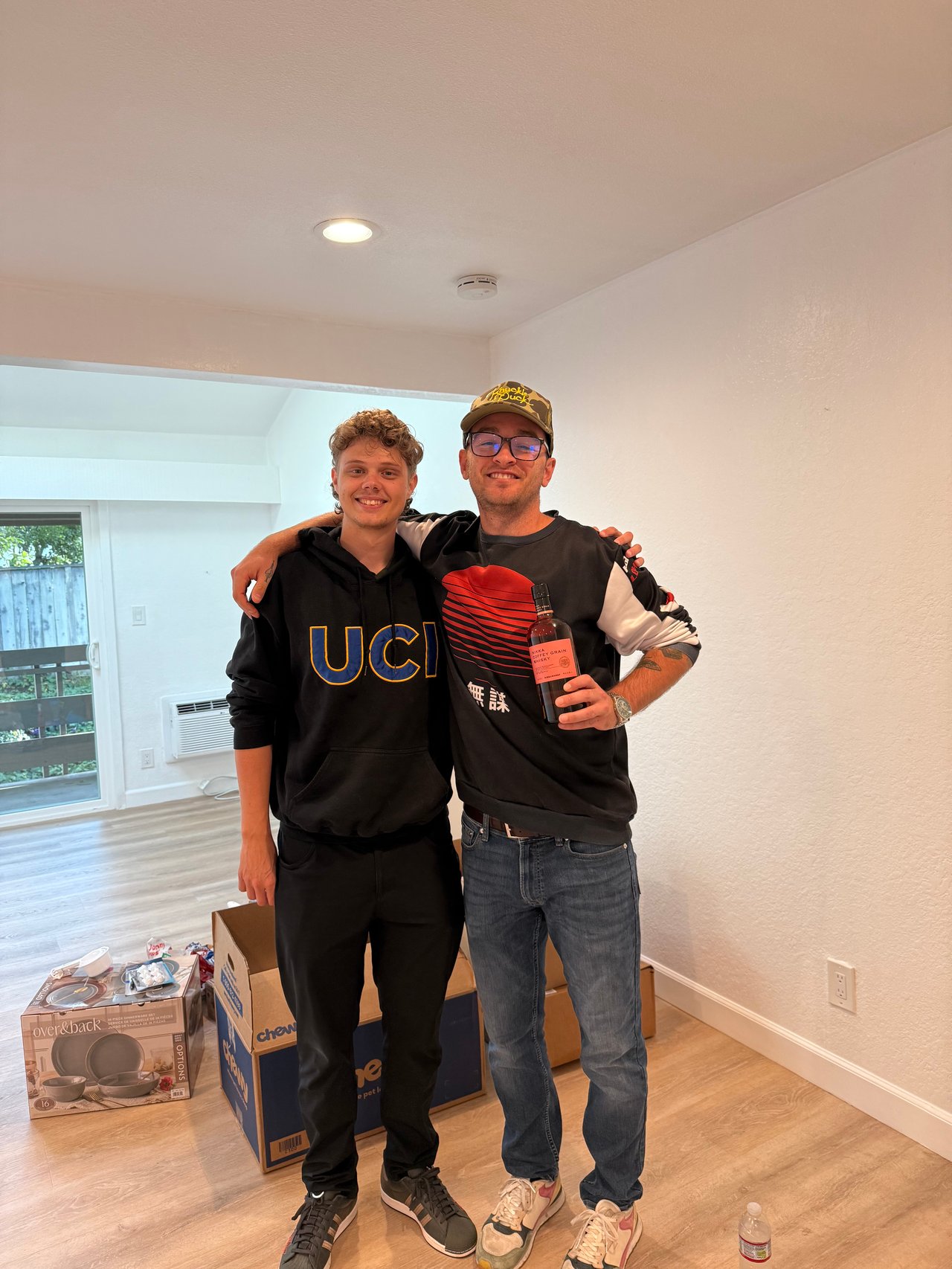Two men pose together in a mostly empty room, surrounded by moving boxes.