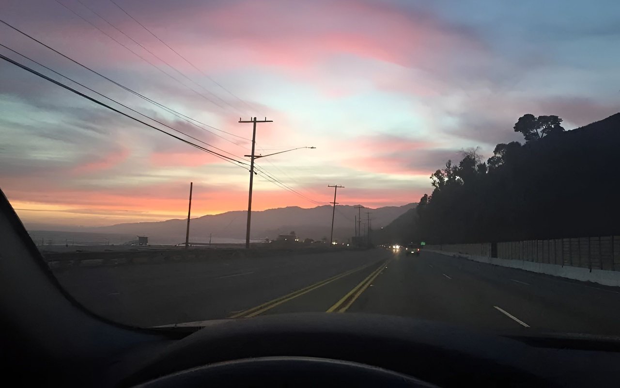 Road toward distant mountains and coastline beneath a pink sunset sky, seen from inside a car.
