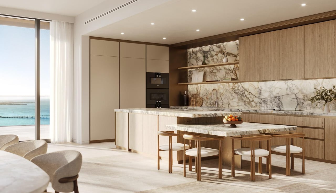 Luxury apartment kitchen with marble counters, pale wood cabinetry, and water views through floor-to-ceiling windows.