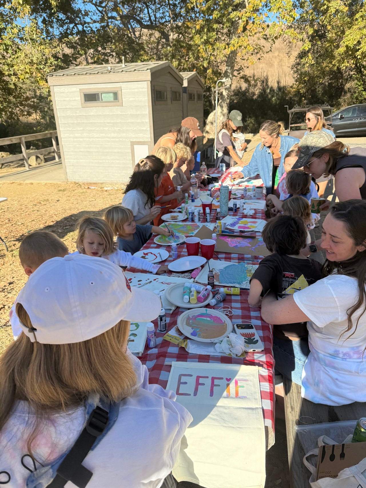 Adults and children paint and craft together at picnic tables beside campground cabins.