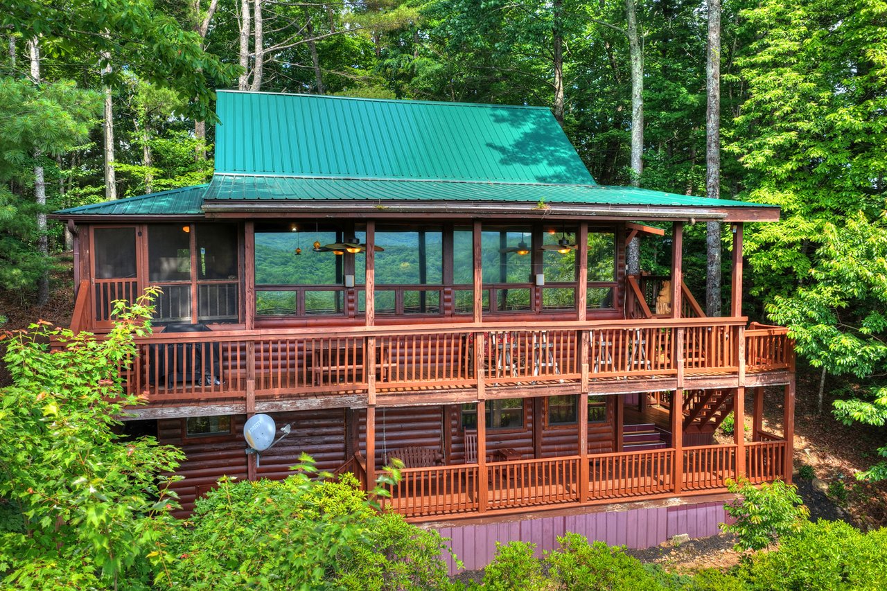 Mountain cabin with a green metal roof, wraparound screened porch, and wooded mountain views.