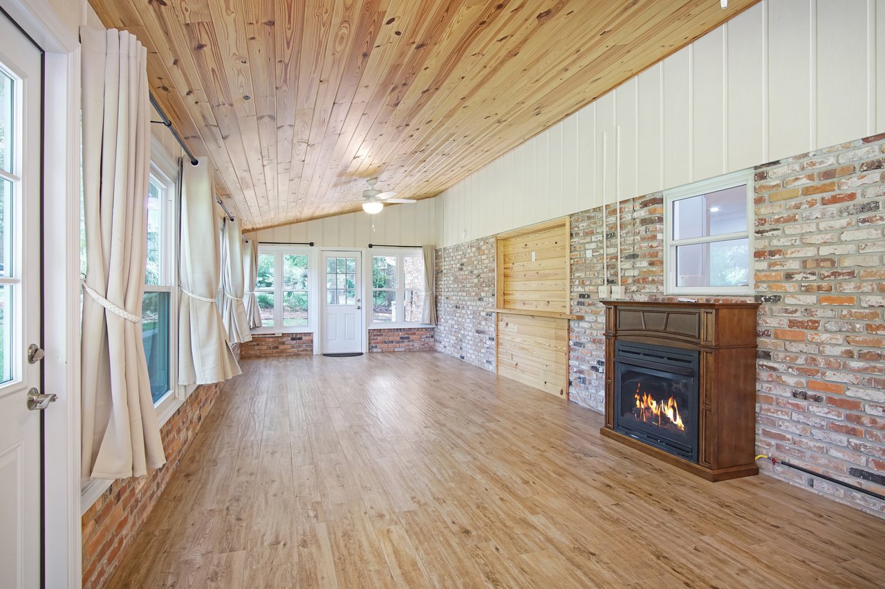 Warm, rustic room with wood ceiling and floor, brick walls, large windows with beige curtains, a fireplace, and white door. Cozy, inviting feel.