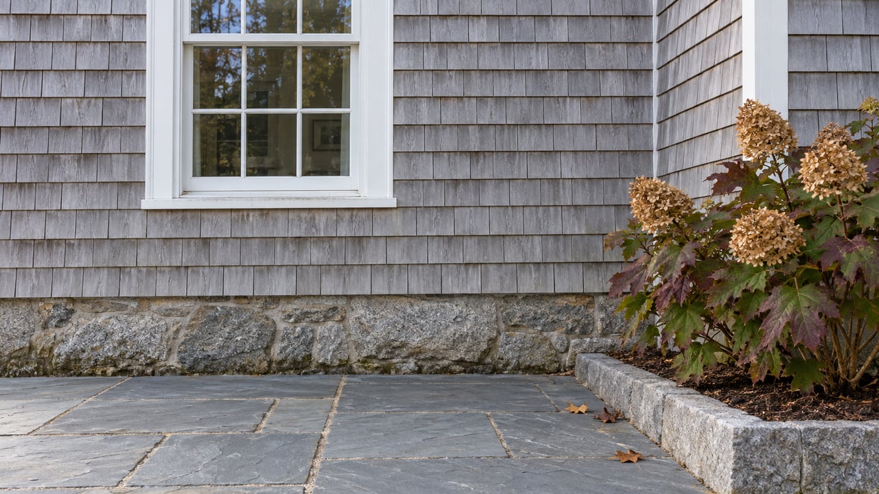 Weathered cedar shingles and a white sash window above a granite foundation and slate terrace, beside an oakleaf hydrangea with autumn foliage.