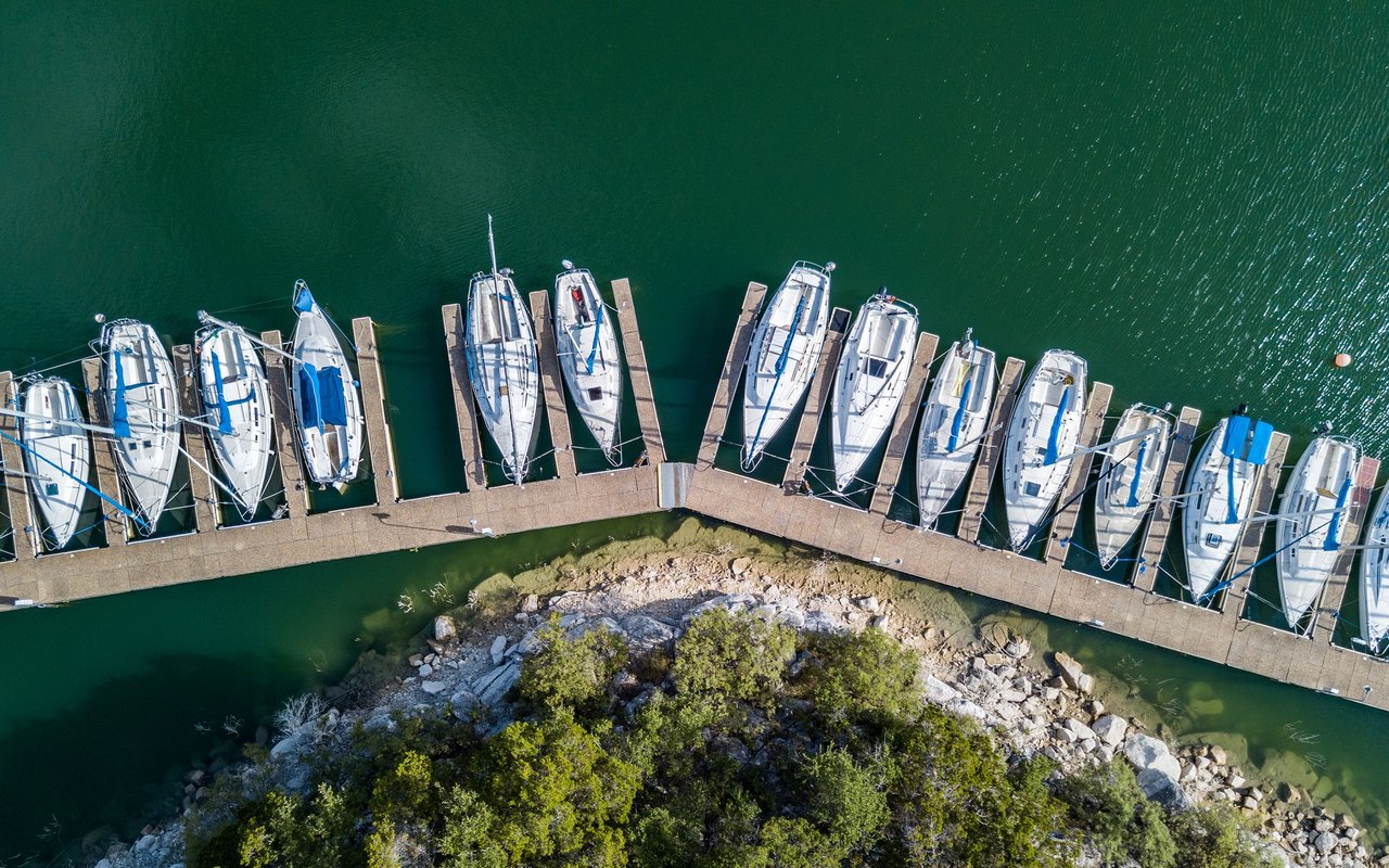 Boats moored along a curved marina dock beside Lakeway’s rocky, tree-lined shore.