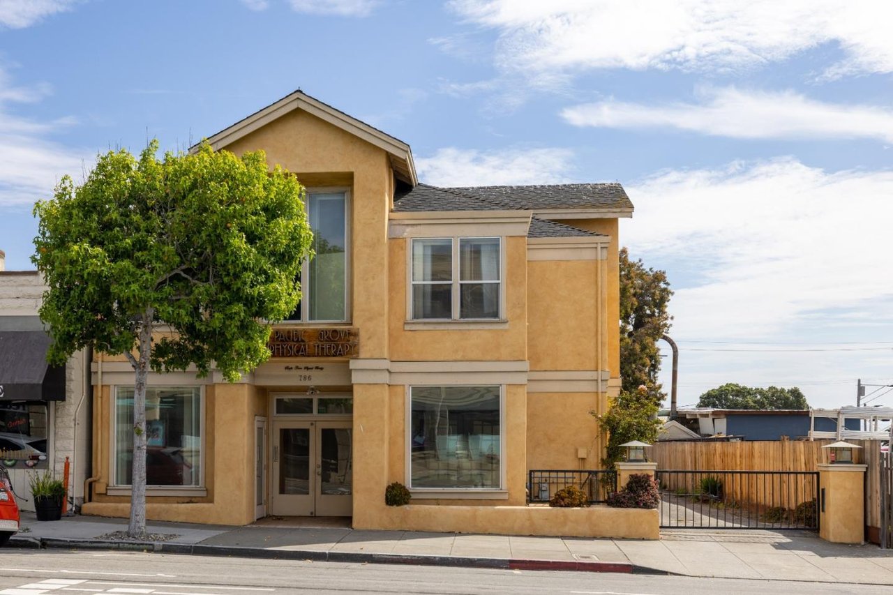 Two-story tan stucco commercial building with a peaked roof, large windows, and a gated side driveway.