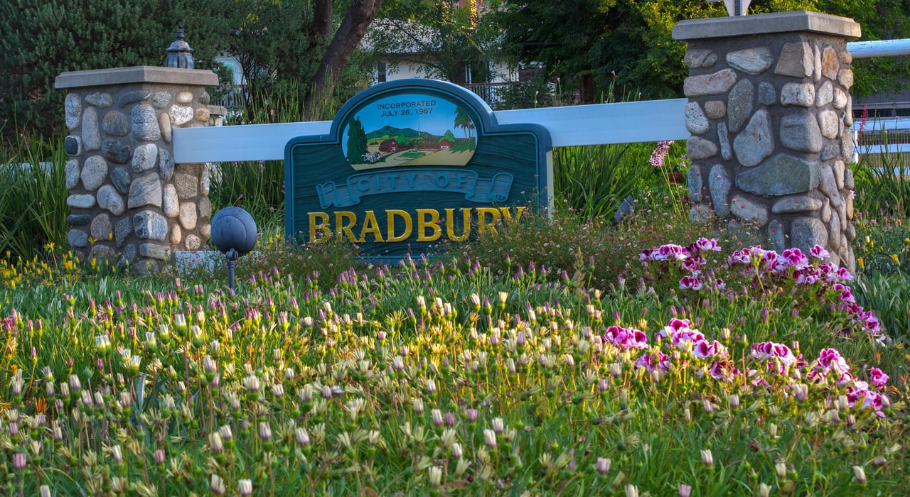 City of Bradbury sign between stone posts and white fencing, surrounded by flowering landscaping