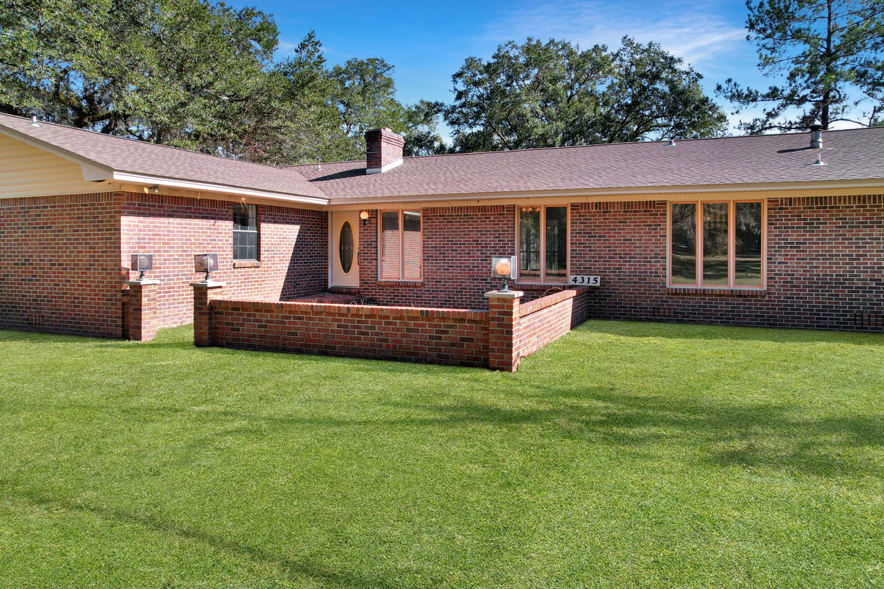 Brick ranch-style house with a brown roof, surrounded by lush green grass. A small brick wall encloses a welcoming entrance. Sky is clear and blue.