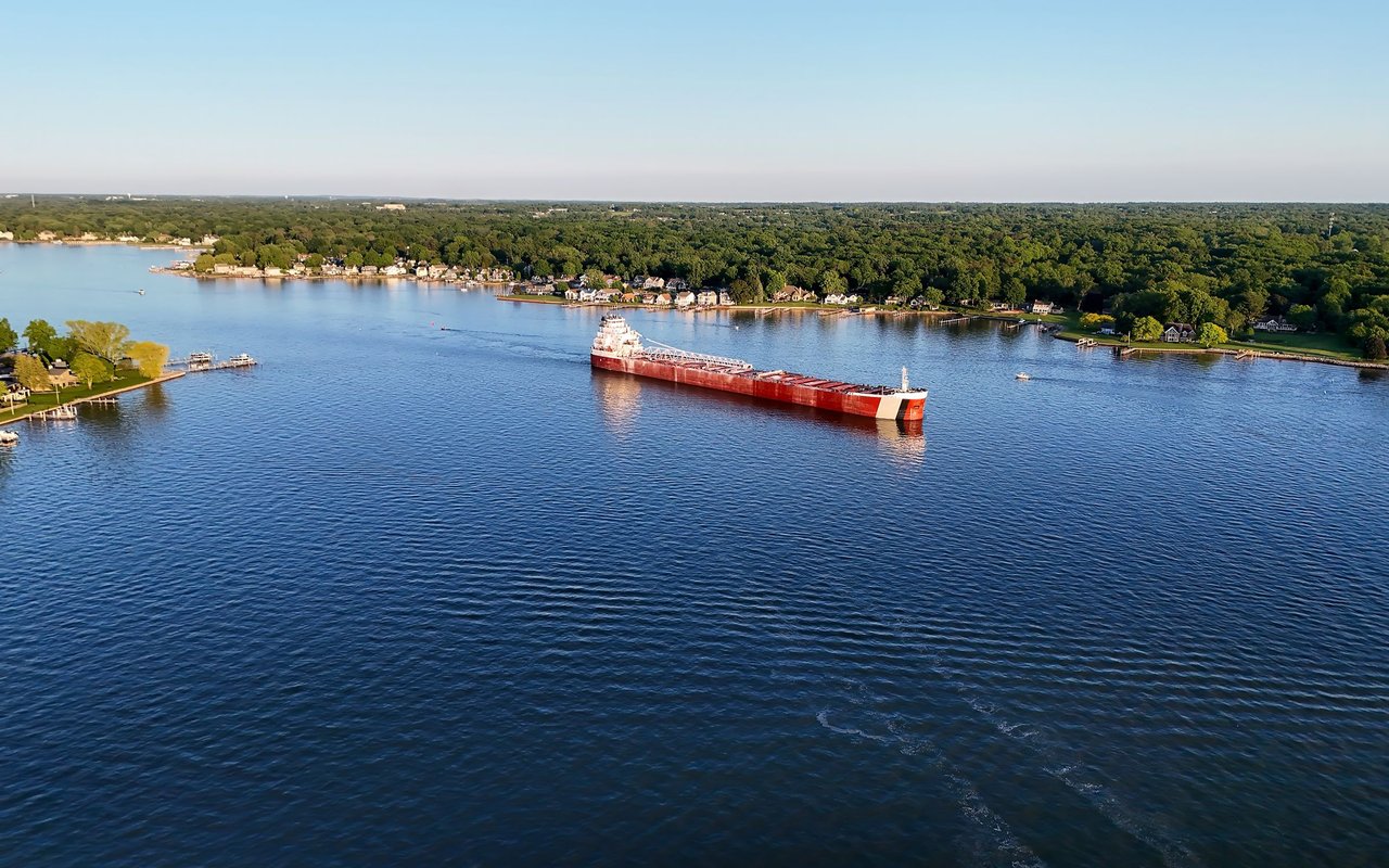 A red-and-white cargo ship crosses a broad river beside a wooded shoreline of homes and docks.